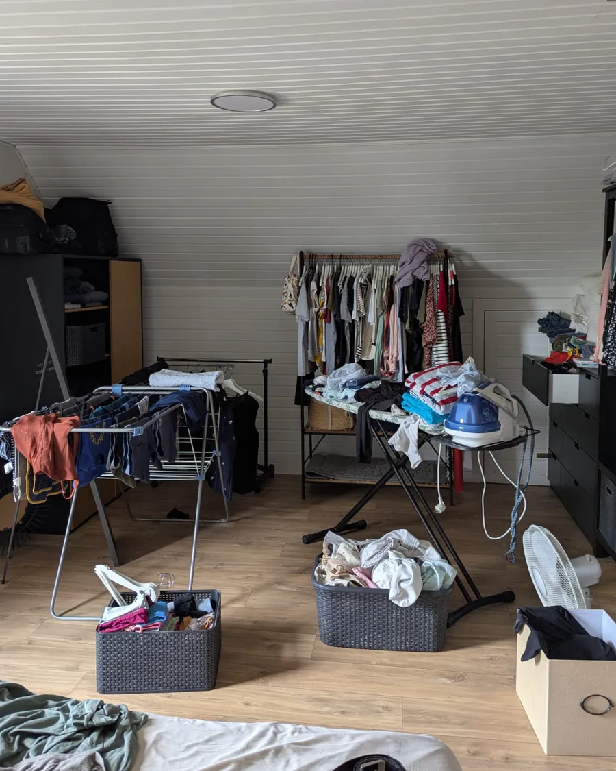 Dressing sous combles avant rangement : étendoirs chargés, planche à repasser, paniers et cartons au milieu du sol